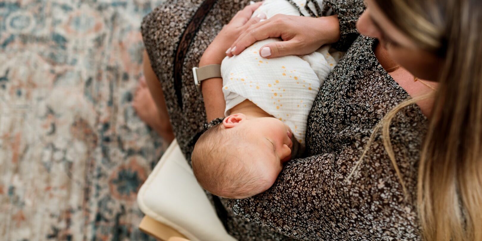 A woman holds and snuggles her sleeping baby. The baby is in a swaddle and is peaceful. The mom is wearing a dark flowered dress and the image is from above.