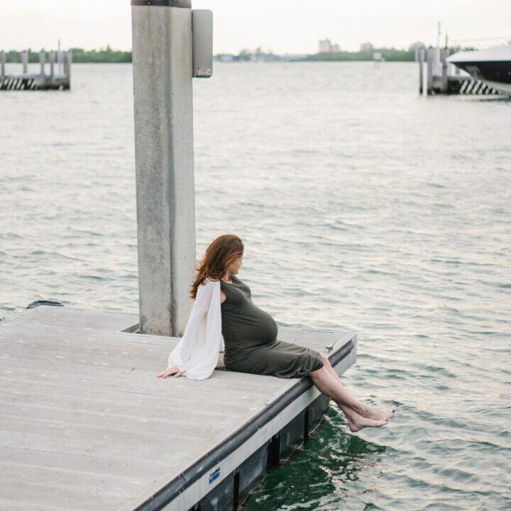 Side view of unrecognizable pregnant future mother in casual clothes enjoying weather while resting on wooden pier in summer